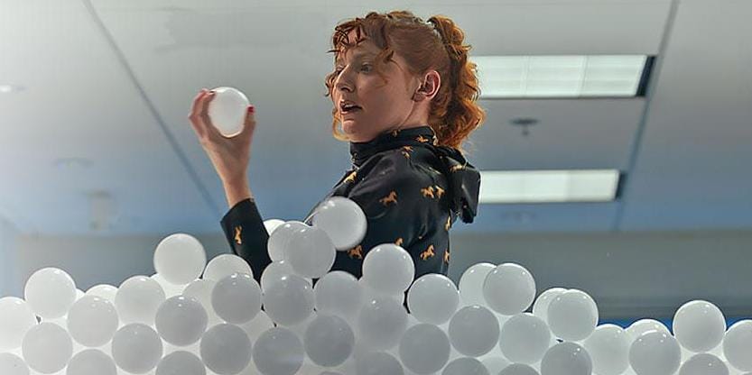 Woman standing in office filled with white balls representing data, holding and curiously looking at a ball