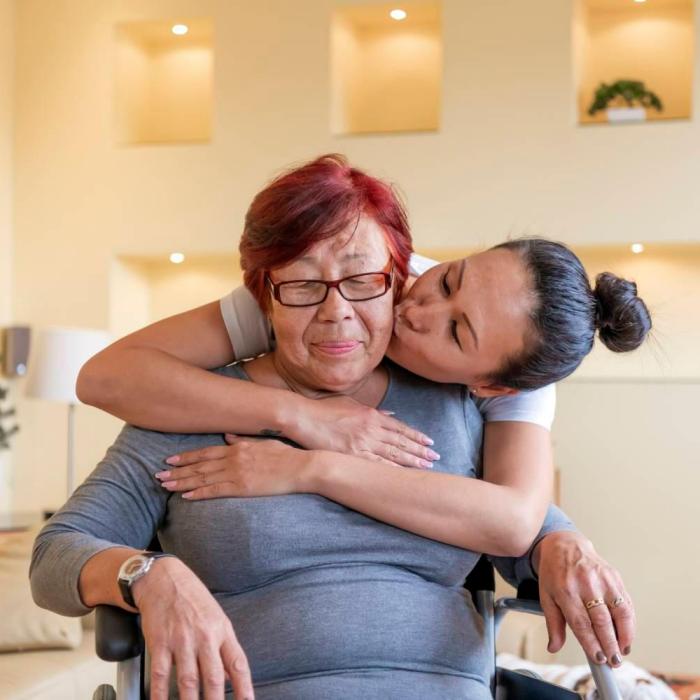 Woman embracing older woman in wheelchair A younger woman embraces an older woman in a wheelchair from behind and kisses her on the cheek.
