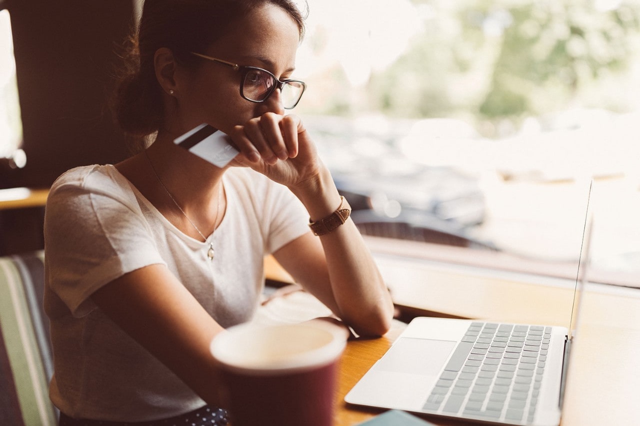 Woman in front of her laptop holding credit card with worried expression on her face