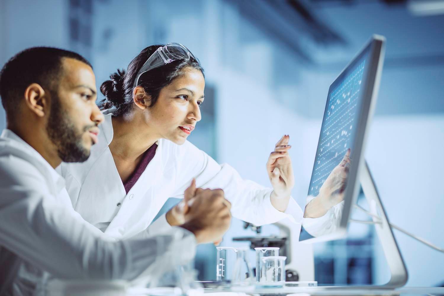 Two scientists in white lab coats reviewing results on a monitor.