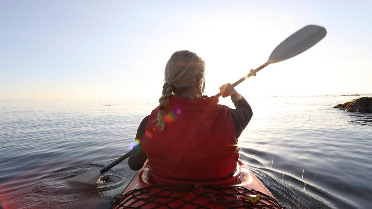 Woman paddles kayak on calm sea, towards sunrise