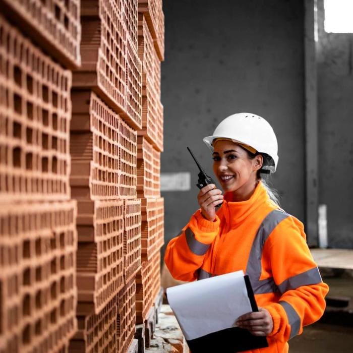 A female worker in a brick factory, wearing an orange jacket and white hardhat, speaks into a walkie talkie.
