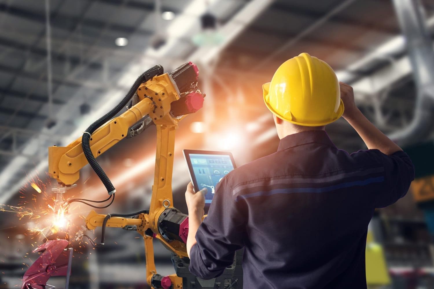 Engineer in yellow hardhat holds tablet computer while watching robotic arm weld metal