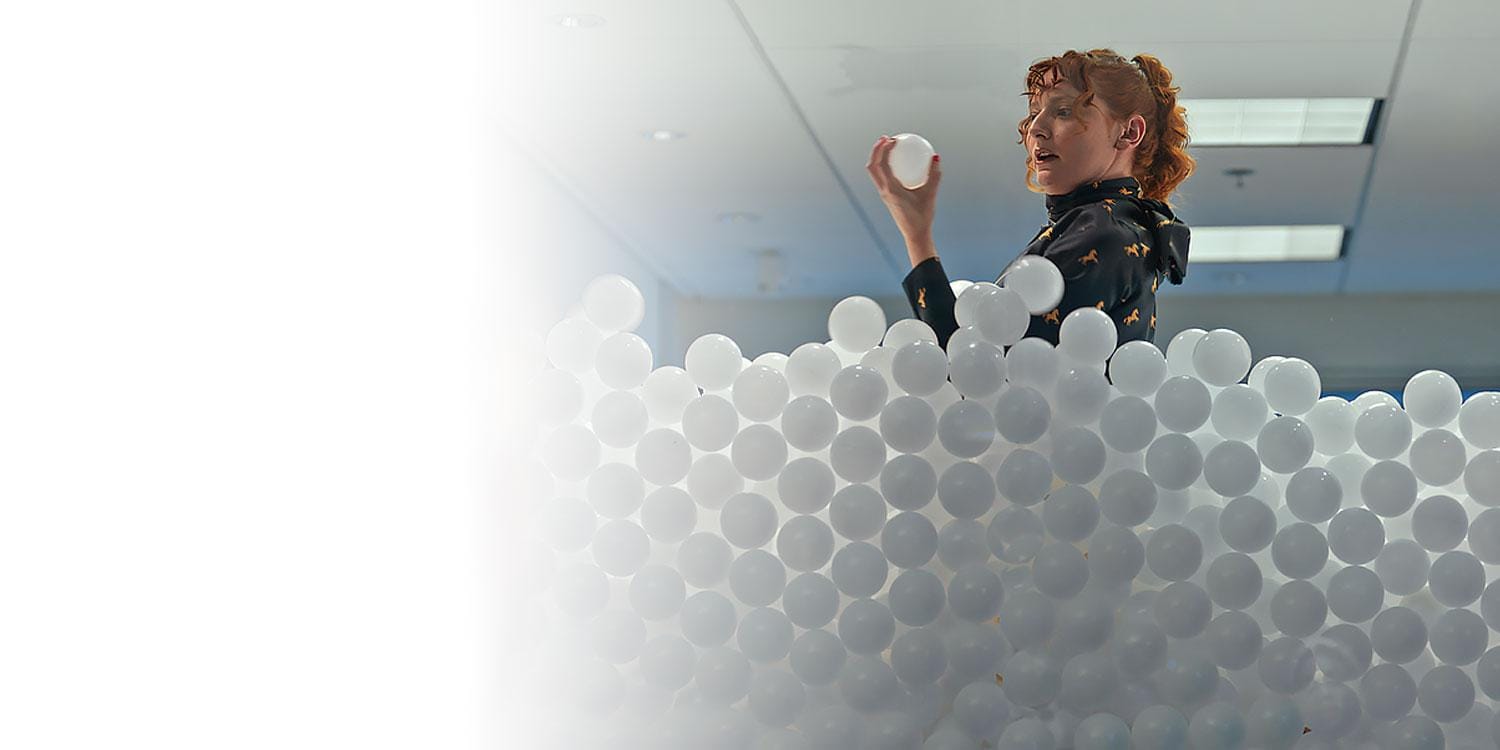 Woman standing in office filled with white balls representing data, holding and curiously looking at a ball
