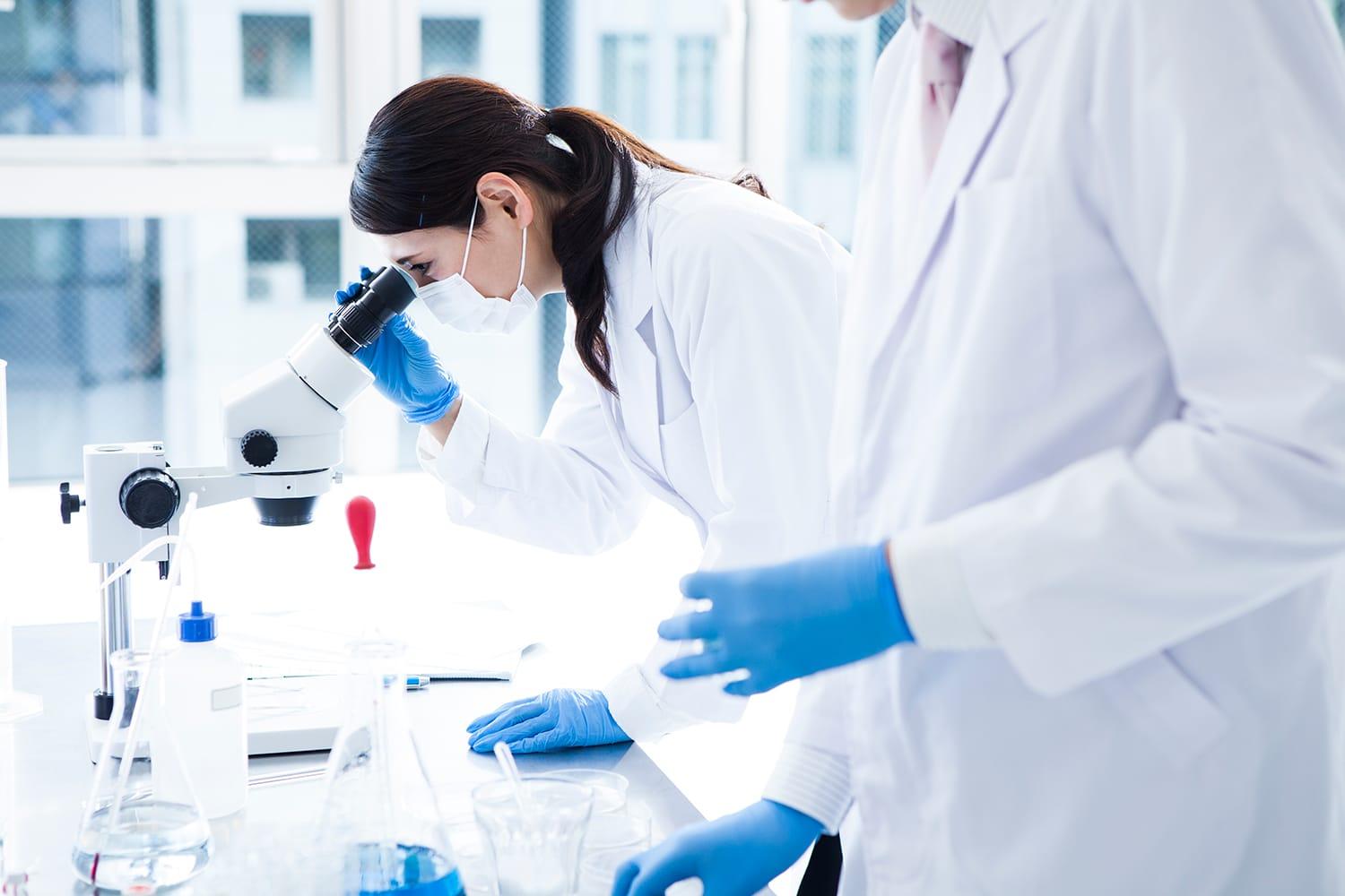 Female clinical research scientist in white lab coat peers into microscope in a lab.