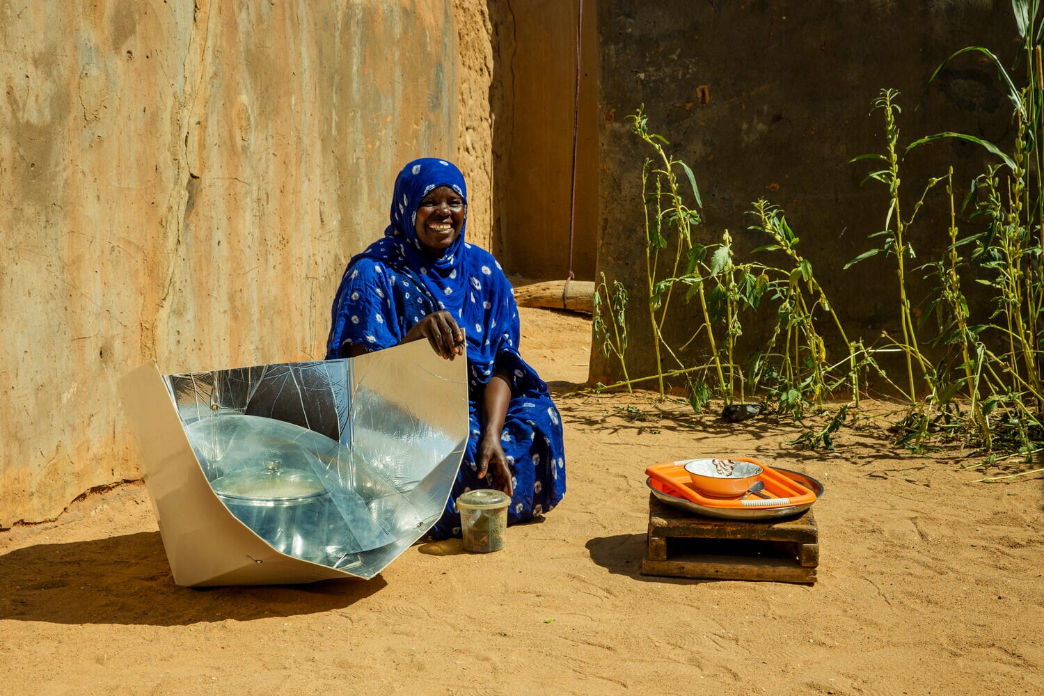 Woman wearing bright blue outift, smiling as she sits beside a solar cookstove