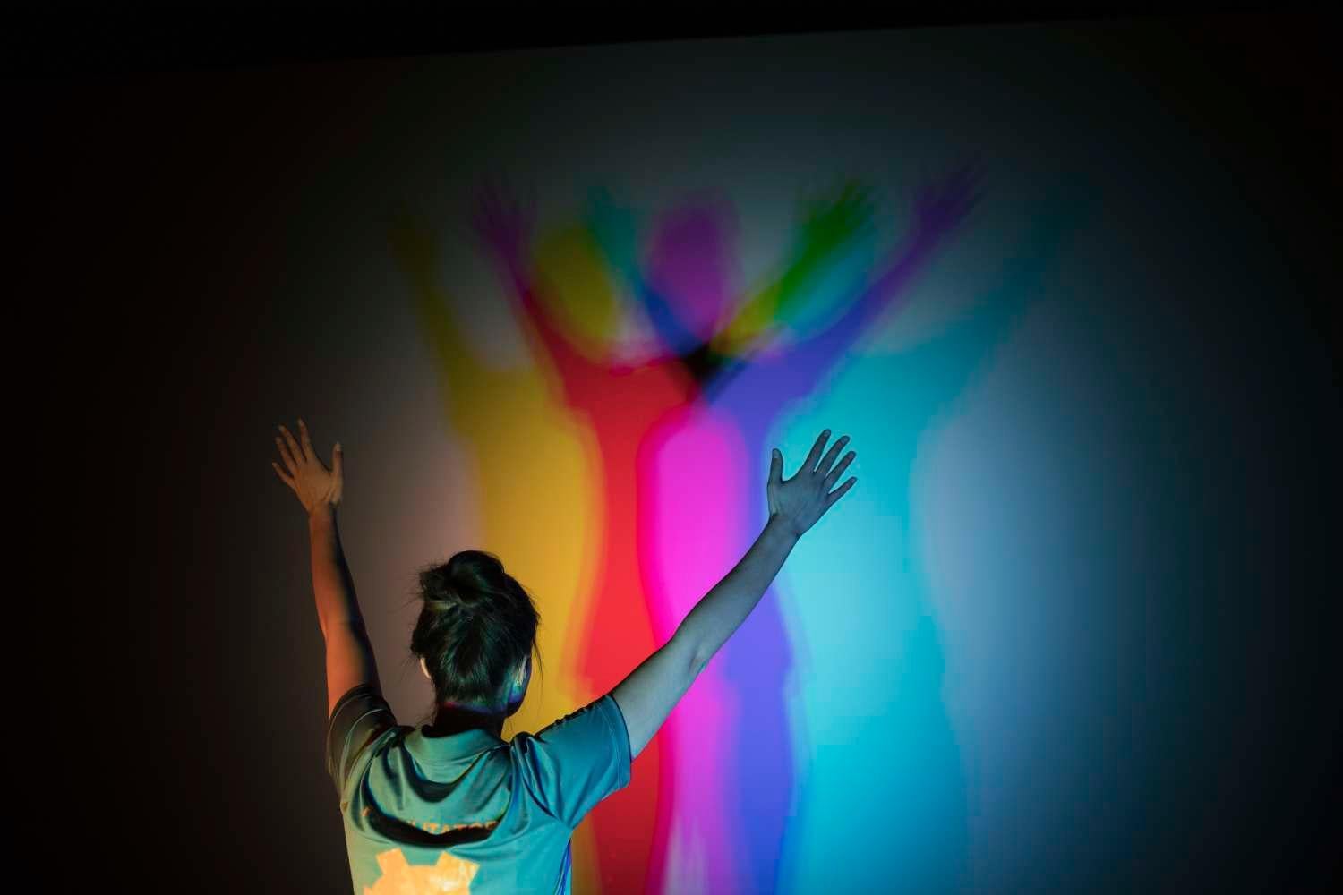 Woman with arms raised views her shadow as a spectrum of colors on wall