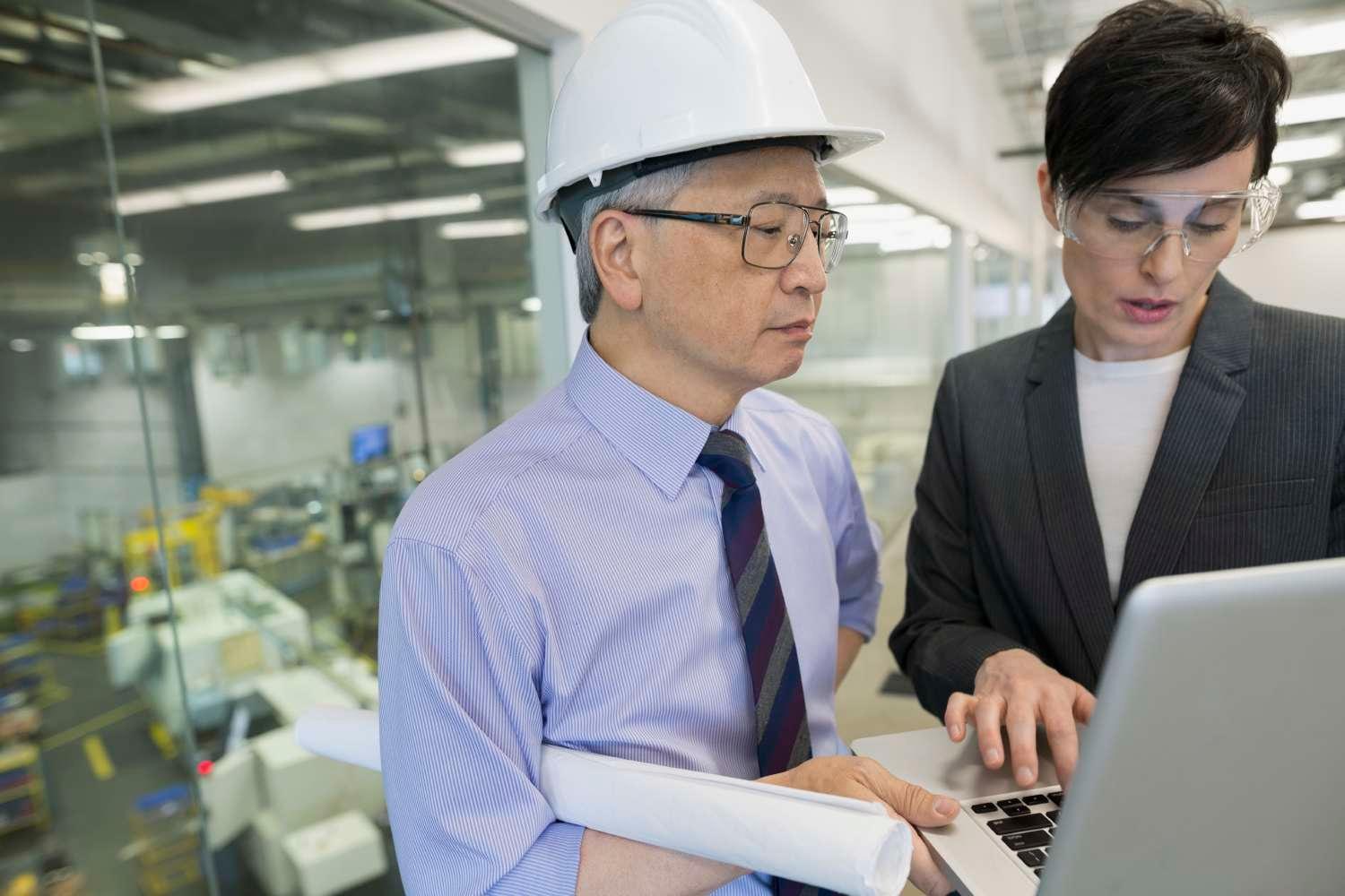 Woman with safety glasses and man wearing a hard hat look at computer in factory hall