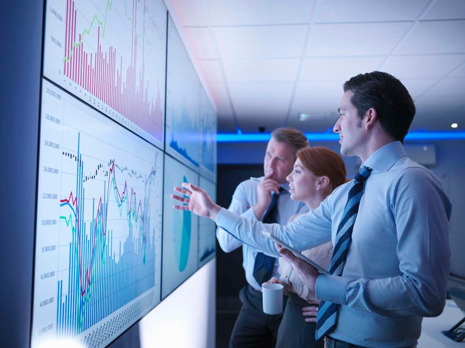 Three businesspeople look at graphs on a large, illuminated screen.