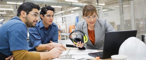 Two men in blue shirts and an executive woman in manufacturing setting looking at computer