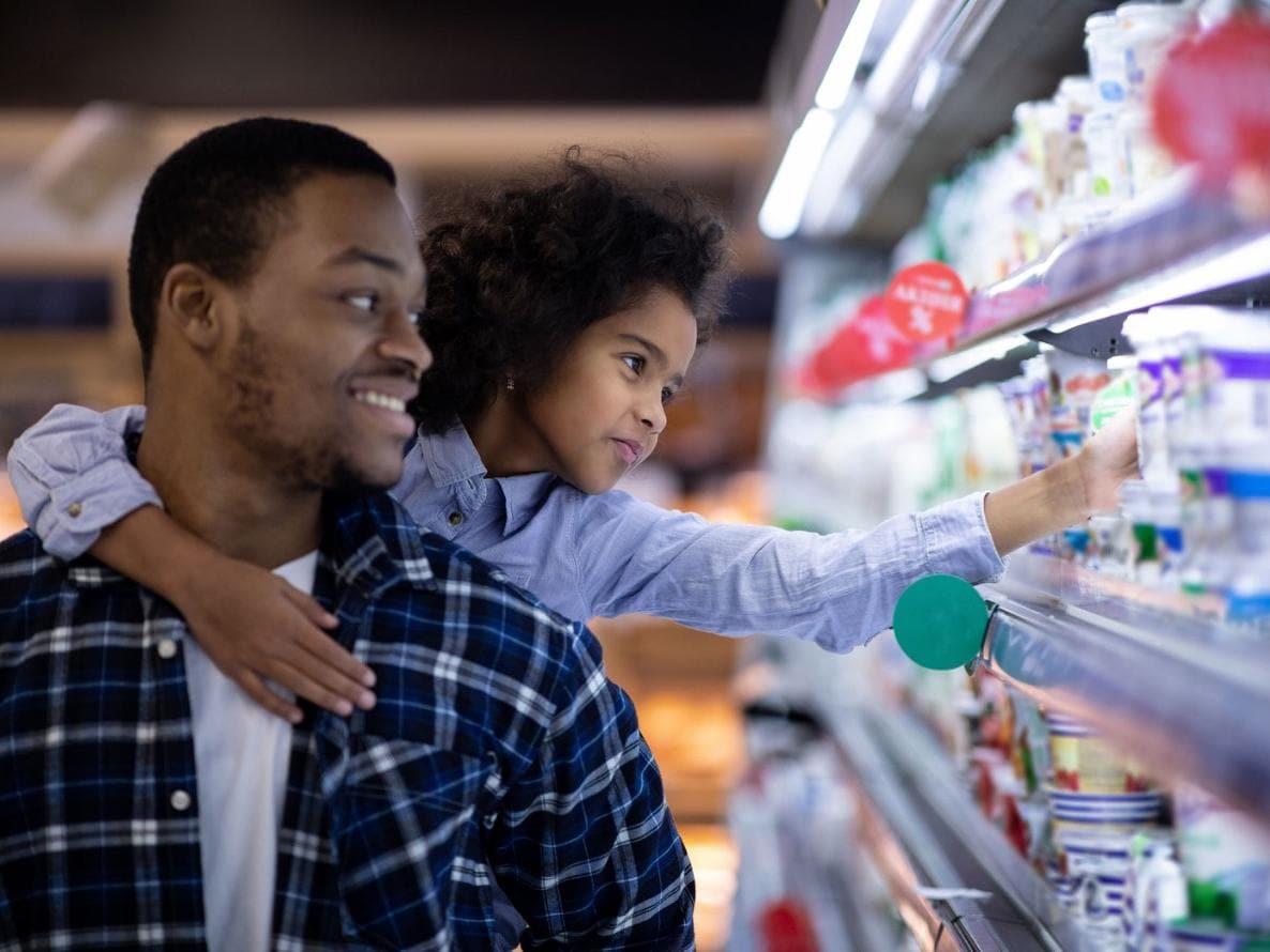 Un hombre sonríe mientras ayuda a su hija a alcanzar los artículos de la estantería de un supermercado