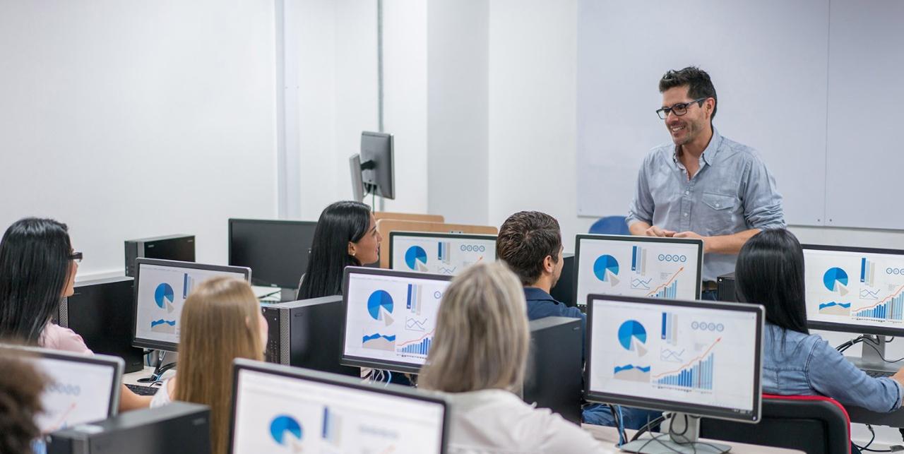 Man standing in front of a class of students sitting behind computer monitors Man standing in front of a class of students sitting behind computer monitors