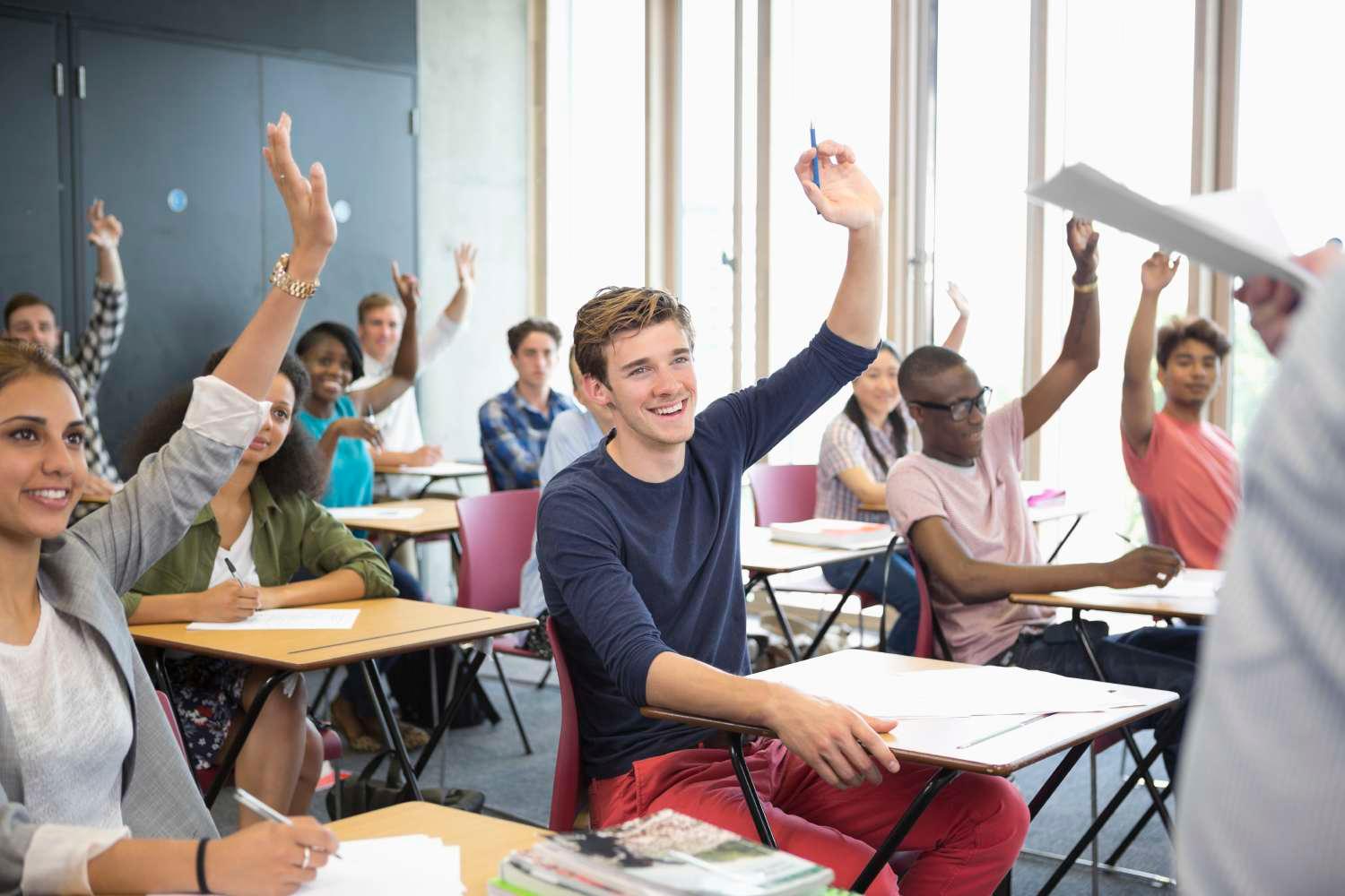 Smiling students sitting at desks in classroom with arms raised