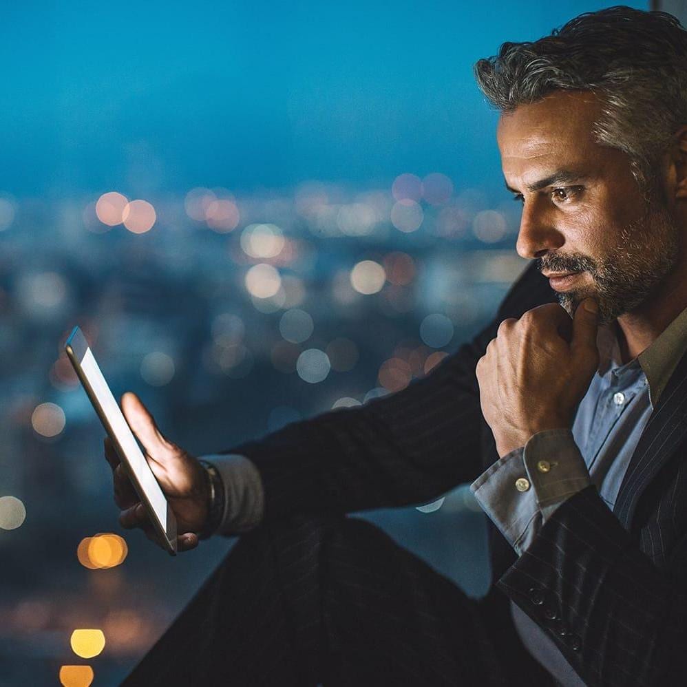 Businessman sitting on a windowsill at night and looking at a digital tablet