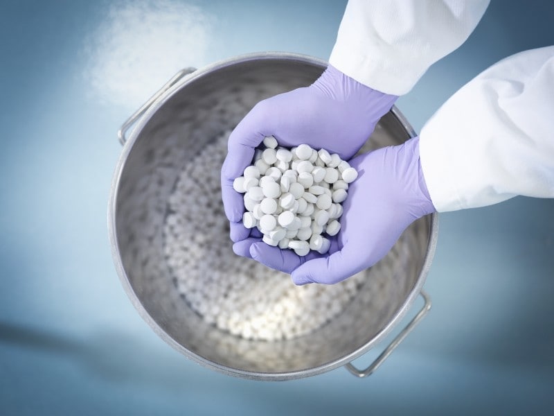 Ręce w rękawiczkach trzymające białe tabletki wyjęte z większego pojemnika wypełnionego większą ilością tych samych tabletek.