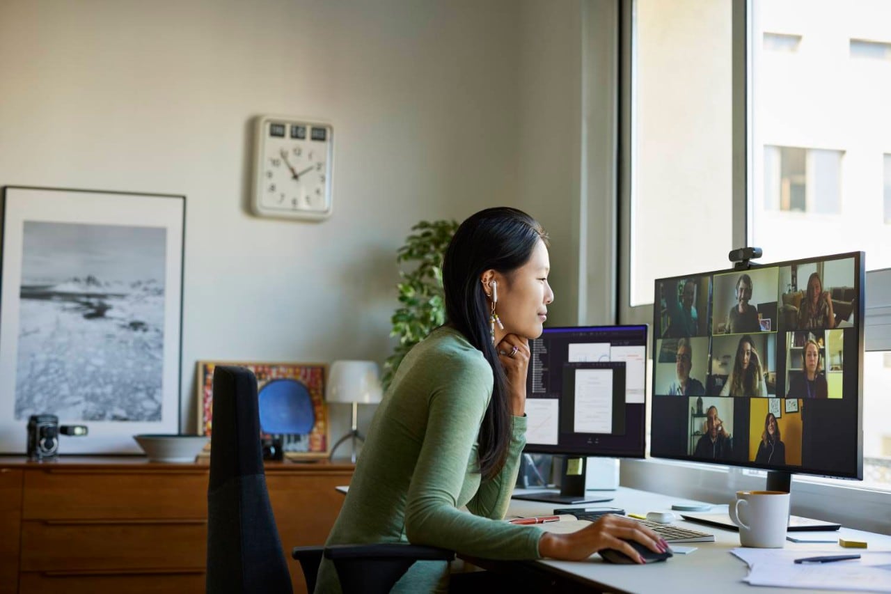 Uma mulher sentada em um escritório, usando fones de ouvido e participando ativamente de uma chamada de videoconferência ao vivo com várias pessoas.