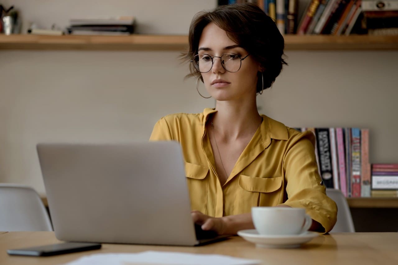 Young attractive woman in eyeglasses dreamily working on laptop with cup of coffee in office