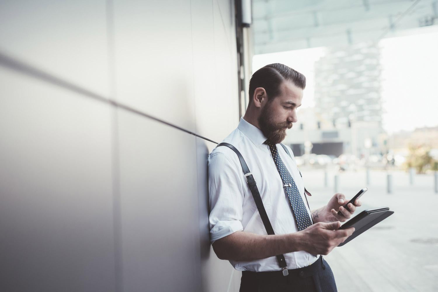 Young businessman leaning on an office wall using smartphone and digital tablet