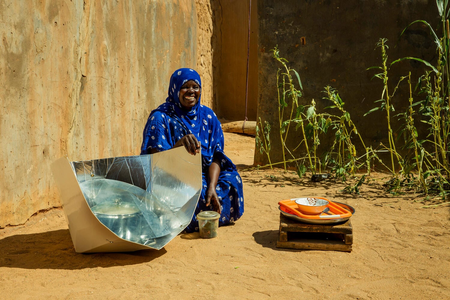 Woman wearing bright blue outift, smiling as she sits beside a solar cookstove
