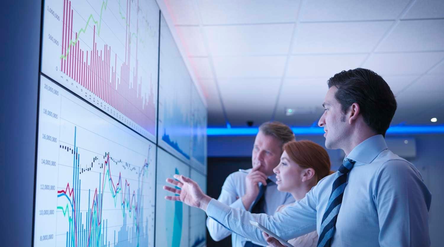 Three business people discuss graphs on screen in meeting room   