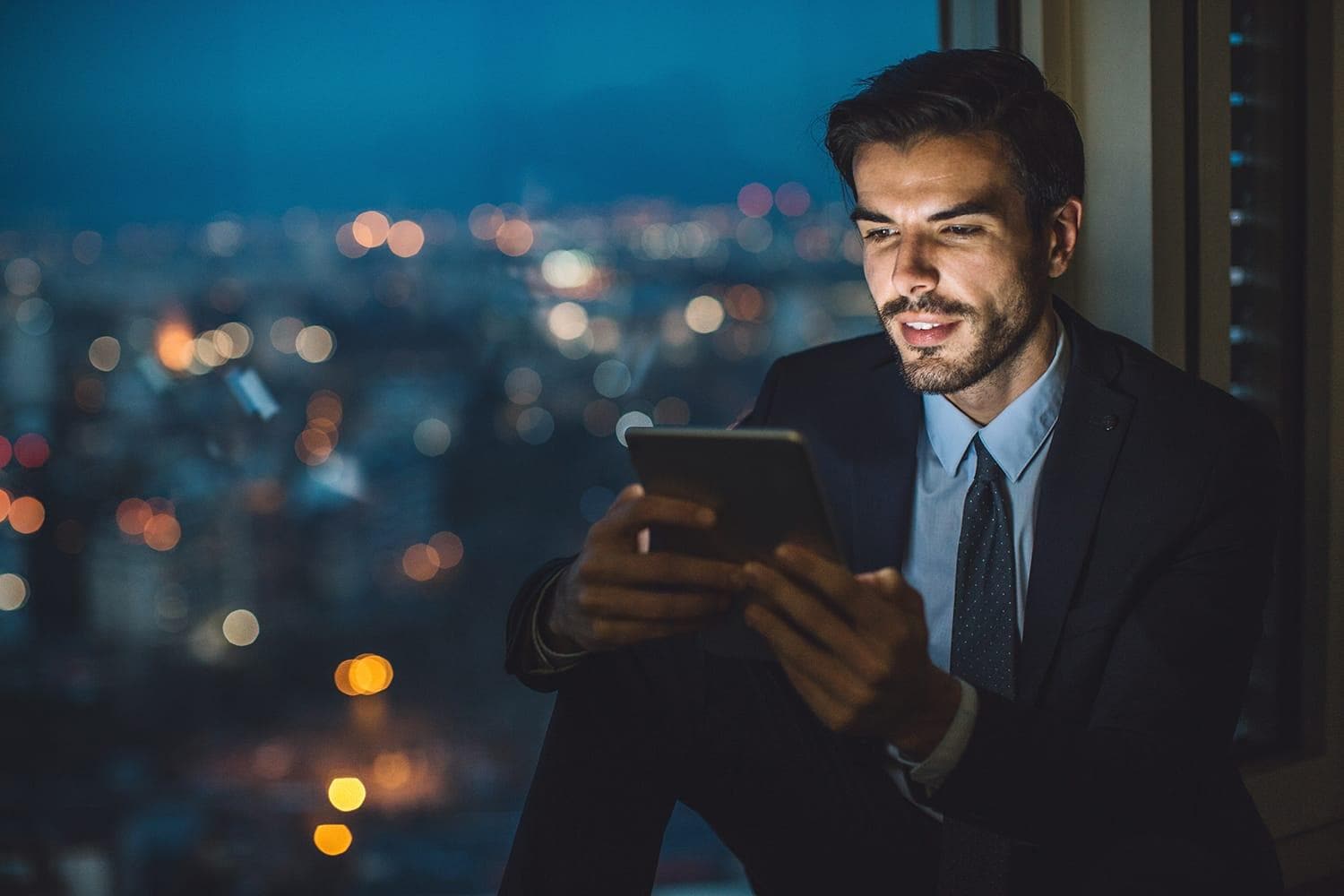 Businessman sitting on a windowsill at night and looking at a digital tablet with cityscape at the background